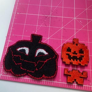 May include: Three Halloween-themed patches. A large black pumpkin patch with a red outline and white eyes. Two smaller orange pixelated pumpkin patches. The patches are on a pink cutting mat.
