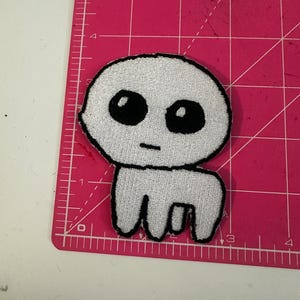 Può includere: Una toppa ricamata bianca raffigurante una creatura dei cartoni animati con grandi occhi neri e una bocca piccola. La creatura ha una testa arrotondata e un corpo con quattro zampe corte. La toppa ha un bordo nero.