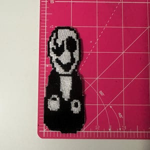 Könnte beinhalten: Ein pixeliger Aufnäher mit einer Figur mit weißem Gesicht und schwarzem Körper. Die Figur hat ein breites Grinsen und große, dunkle Augen. Der Aufnäher befindet sich auf einer rosa Schneidematte mit Zentimetermaßen.