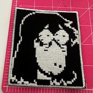 Puede incluir: Parche bordado cuadrado en blanco y negro con un diseño de rostro pixelado. La imagen está creada con hilo blanco sobre un fondo negro, en un estilo de bloques. El parche tiene un borde blanco.