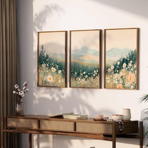 May include: Three framed landscape art prints. Each print features a watercolor painting of a mountain scene with a field of daisies in the foreground. The frames are a warm brown color, and the overall color palette is soft and natural.