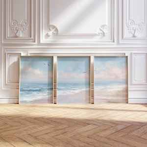 May include: Three framed landscape paintings depicting a serene beach scene with a blue ocean, pink-tinged clouds, and flying birds. The paintings are displayed against a white wall with decorative molding, and the floor is made of wooden planks.