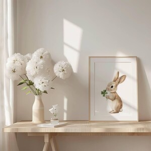 May include: A framed print of a brown rabbit holding a green four-leaf clover. The print is on a light wood table with a vase of white flowers.