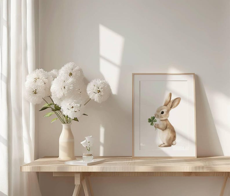 May include: A framed print of a brown rabbit holding a green four-leaf clover. The print is on a light wood table with a vase of white flowers.