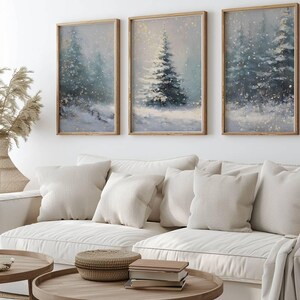 May include: Three framed winter landscape paintings with snow-covered evergreen trees. The artwork has a soft, muted color palette of blues, greens, and whites, with a touch of gold. The paintings are displayed above a white sofa.
