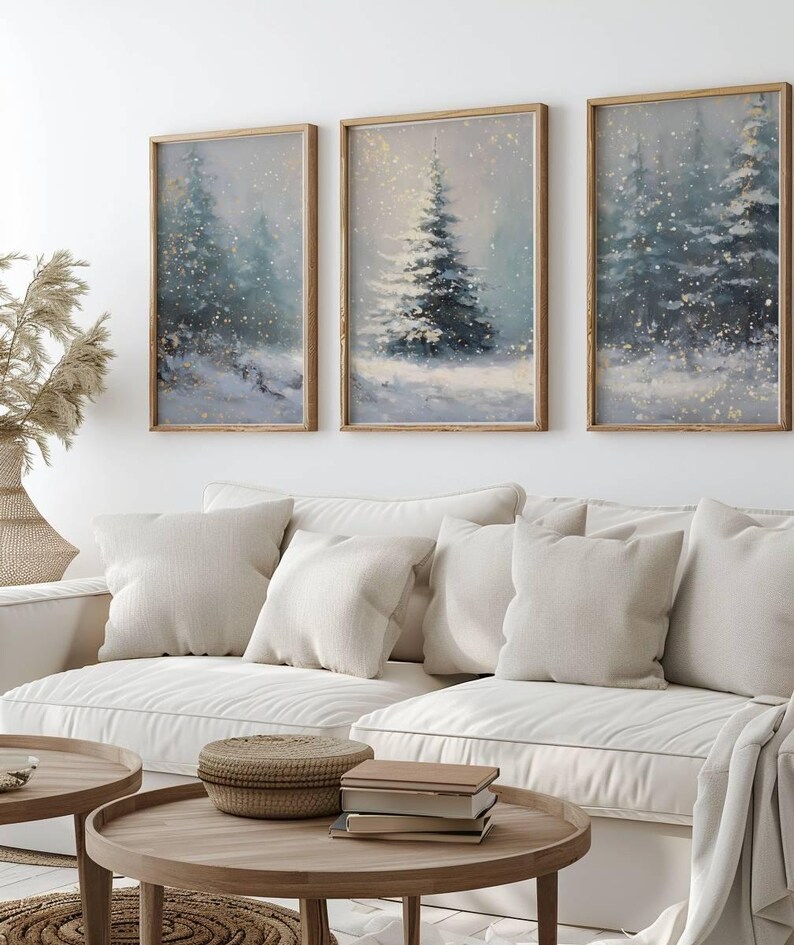 May include: Three framed winter landscape paintings with snow-covered evergreen trees. The artwork has a soft, muted color palette of blues, greens, and whites, with a touch of gold. The paintings are displayed above a white sofa.