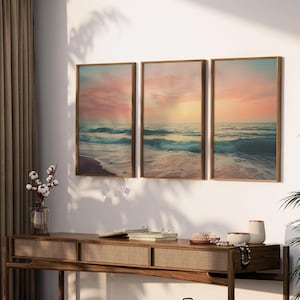 Puede incluir: Tres impresiones enmarcadas de una escena de playa con un cielo de puesta de sol y olas rompiendo en la orilla. Las impresiones est&aacute;n colgadas en una pared blanca sobre una mesa consola de madera.