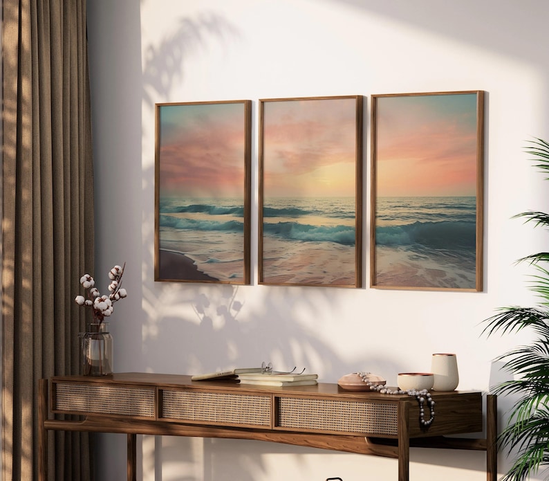 Puede incluir: Tres impresiones enmarcadas de una escena de playa con un cielo de puesta de sol y olas rompiendo en la orilla. Las impresiones est&aacute;n colgadas en una pared blanca sobre una mesa consola de madera.