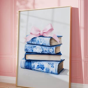 May include: A framed artwork featuring a stack of blue and white floral-patterned books tied with a pink bow. The books have gold-colored pages and are set against a white background. The frame is gold-colored.