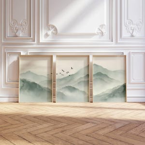 Peut inclure: Trois impressions d'art encadrées représentant un paysage de montagne brumeux dans des tons de vert et de gris. L'œuvre comprend des oiseaux en vol et est exposée sur un sol en bois contre un mur blanc.