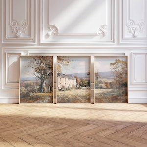 May include: Three framed landscape paintings showing a rural scene with a house, trees, and fields. The artwork uses muted tones of brown, beige, and green, with a light-coloured house as the focal point. The paintings are displayed against a white wall with decorative moulding.