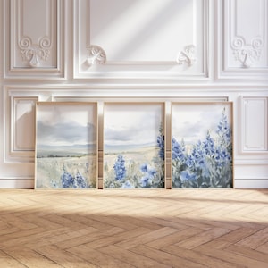 May include: Three framed watercolor paintings depicting a field of blue wildflowers against a cloudy sky. The paintings are in gold frames and are hung on a white wall.