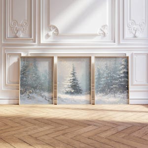 May include: Three framed winter landscape paintings. Each features a snow-covered evergreen tree in a snowy forest scene. The artwork has a soft, muted color palette with gold accents, set against a white wall and a wooden floor.