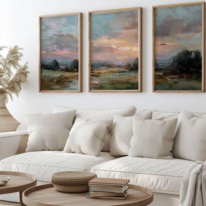 May include: Three framed landscape paintings with a sunset sky in shades of pink, orange, and blue. The artwork is displayed above a white sofa with beige pillows. Wooden coffee tables and a woven basket are in the foreground.