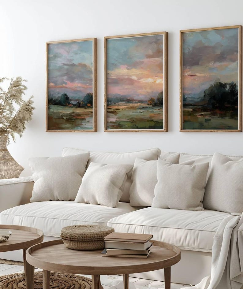 May include: Three framed landscape paintings with a sunset sky in shades of pink, orange, and blue. The artwork is displayed above a white sofa with beige pillows. Wooden coffee tables and a woven basket are in the foreground.