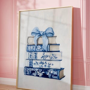 May include: A framed artwork showing a stack of books with blue and white floral patterns. A light blue bow is tied on top. The artwork is set against a pink wall and wooden floor.