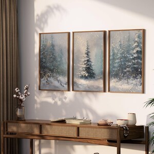 May include: Three framed winter landscape paintings with snow-covered evergreen trees. The artwork features a muted color palette of blues, whites, and golds. The paintings are displayed above a wooden console table with decorative items.