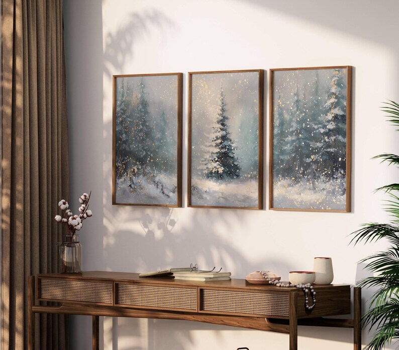 May include: Three framed winter landscape paintings with snow-covered evergreen trees. The artwork features a muted color palette of blues, whites, and golds. The paintings are displayed above a wooden console table with decorative items.