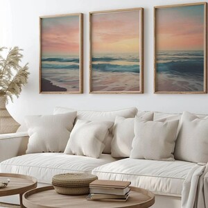 Puede incluir: Tres impresiones enmarcadas de una escena de playa con olas rompiendo en la orilla al atardecer. Las impresiones est&aacute;n en un marco de madera clara y est&aacute;n colgadas en una pared blanca sobre un sof&aacute; blanco con almohadas.