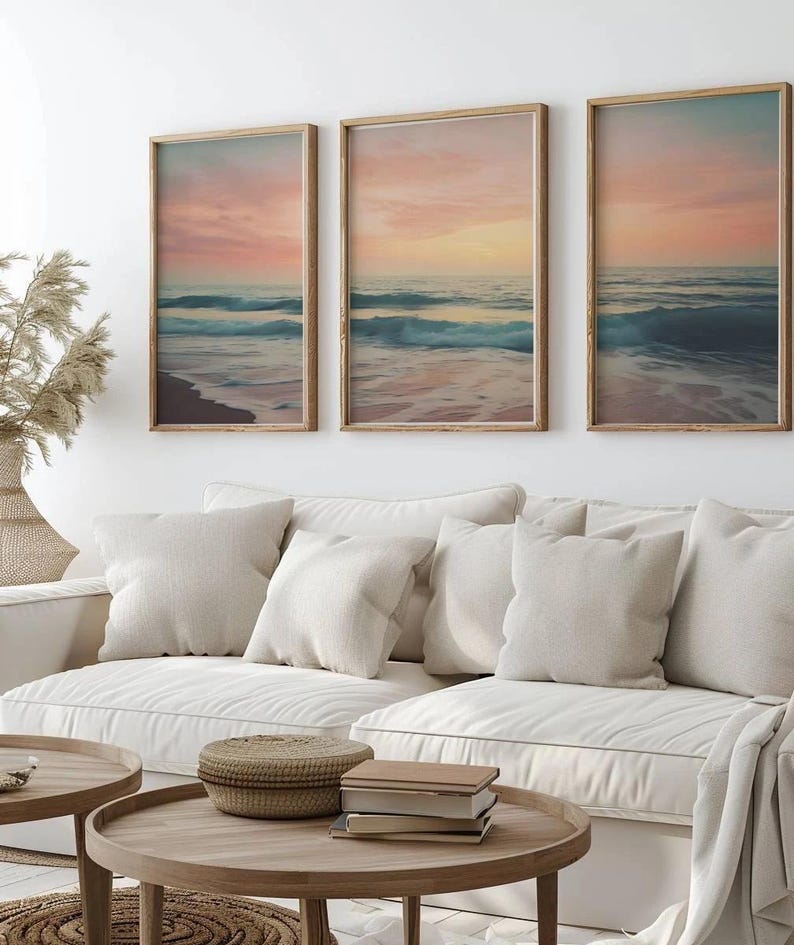 Puede incluir: Tres impresiones enmarcadas de una escena de playa con olas rompiendo en la orilla al atardecer. Las impresiones est&aacute;n en un marco de madera clara y est&aacute;n colgadas en una pared blanca sobre un sof&aacute; blanco con almohadas.