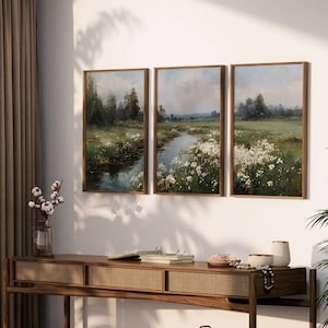 Könnte beinhalten: Drei gerahmte Landschaftsbilder, die eine ruhige Flussszene mit weißen Wildblumen und üppigem Grün zeigen. Die Gemälde haben braune Holzrahmen und werden über einer Holzkonsolentisch ausgestellt.