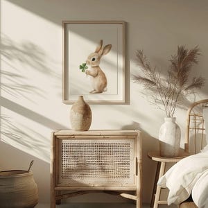 May include: A framed print of a brown rabbit holding a green four-leaf clover. The print is hanging on a white wall above a wooden cabinet with a woven rattan front.