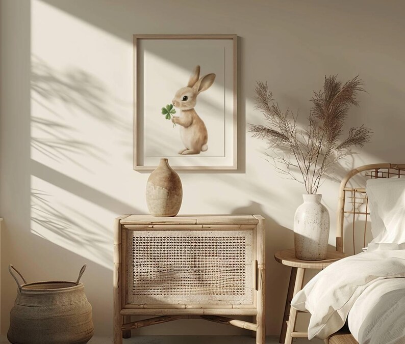 May include: A framed print of a brown rabbit holding a green four-leaf clover. The print is hanging on a white wall above a wooden cabinet with a woven rattan front.