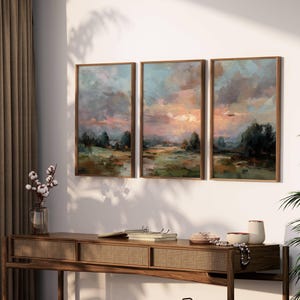 May include: Three framed landscape paintings with a sunset scene in shades of blue, pink, and green. The artwork is displayed above a wooden console table with a woven front. A vase of cotton stems and decorative items are on the table.