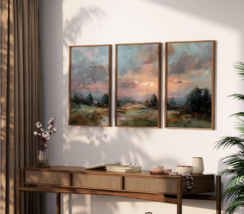 May include: Three framed landscape paintings with a sunset scene in shades of blue, pink, and green. The artwork is displayed above a wooden console table with a woven front. A vase of cotton stems and decorative items are on the table.