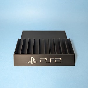 May include: Black plastic storage case with slots for video game cartridges. The case has the text "PS2" printed on the front.
