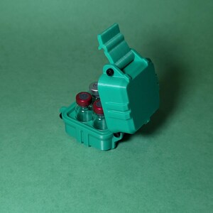 Puede incluir: Una caja de pl&aacute;stico verde azulado con una tapa con bisagras, dise&ntilde;ada para contener peque&ntilde;os viales. La caja est&aacute; abierta, revelando cuatro viales de vidrio transparente con tapas rojas.