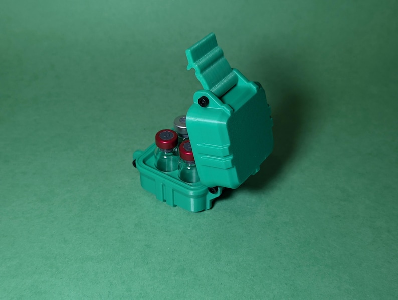 Puede incluir: Una caja de pl&aacute;stico verde azulado con una tapa con bisagras, dise&ntilde;ada para contener peque&ntilde;os viales. La caja est&aacute; abierta, revelando cuatro viales de vidrio transparente con tapas rojas.