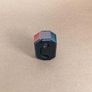 May include: A black and red 3D printed object with a textured surface and two small holes. The object is shaped like a cube with rounded edges.