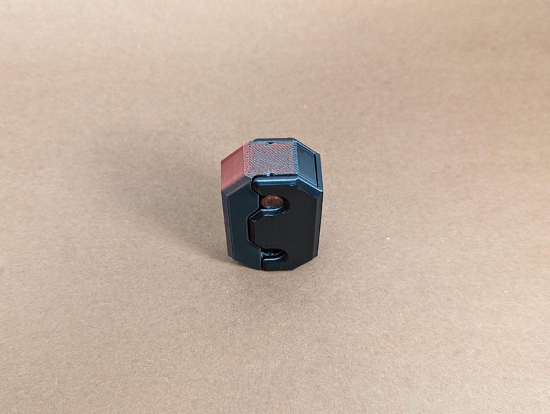 May include: A black and red 3D printed object with a textured surface and two small holes. The object is shaped like a cube with rounded edges.