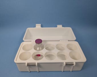 May include: White plastic box with a hinged lid and a handle. The box has six compartments and a single clear glass vial with a purple cap is in one of the compartments.