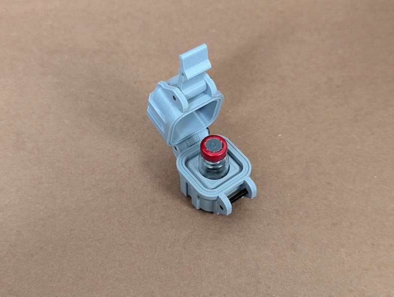May include: A small grey plastic container with a hinged lid, open to reveal a clear glass vial with a red cap. The vial is inside the container.