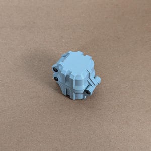 May include: A small, light blue, plastic box with a square shape and a textured surface. The box has a hinged lid and several small, black, plastic tabs.