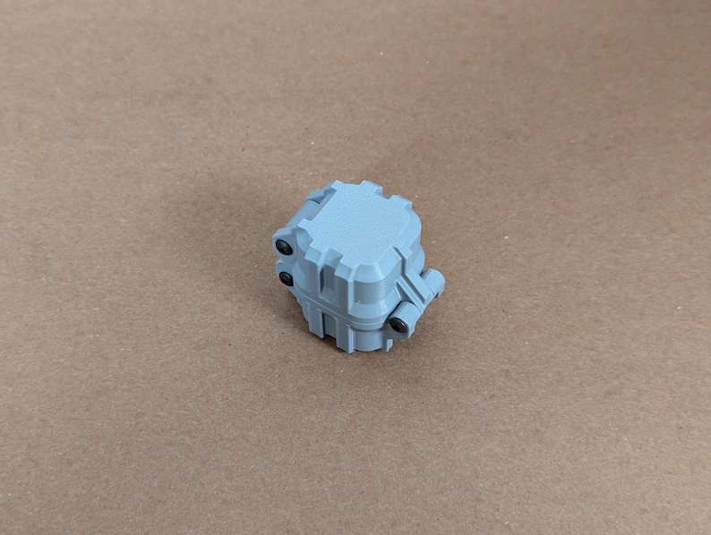 May include: A small, light blue, plastic box with a square shape and a textured surface. The box has a hinged lid and several small, black, plastic tabs.