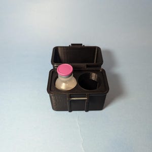 May include: A black plastic case with a hinged lid, open to reveal a small glass vial with a pink cap and a round, empty space.
