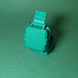 Puede incluir: Una caja de pl&aacute;stico verde azulado con una tapa con bisagras. La caja est&aacute; cerrada y la tapa est&aacute; ligeramente entreabierta. La caja es texturizada y tiene un acabado mate.