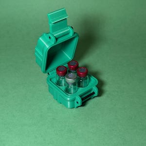 Puede incluir: Una caja de pl&aacute;stico verde con una tapa con bisagras, abierta para revelar seis peque&ntilde;os viales de vidrio con tapas rojas. Los viales est&aacute;n dispuestos en una cuadr&iacute;cula dentro de la caja.