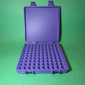 May include: A purple plastic case with a hinged lid and a grid of 64 round compartments. The case is designed to hold small items, such as pills, beads, or other small objects.