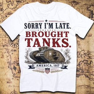 May include: A white t-shirt featuring the text SORRY I'M LATE. BROUGHT TANKS. in dark blue and red, with a graphic of a brown military tank, the text AMERICA, 1917 on a blue banner, and a shield emblem with red and white stripes and blue stars.