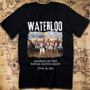 May include: A black short-sleeved t-shirt featuring the text WATERLOO at the top. Below is a colorful painting of soldiers on horseback in battle. The text below the image reads CHARGE OF THE ROYAL SCOTS GREYS and the date June 18, 1815.