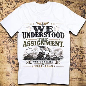 May include: A white t-shirt featuring a graphic print with the text WE UNDERSTOOD THE ASSIGNMENT. The design includes an illustration of soldiers raising the United States flag, a tank, airplanes, and the text UNITED STATES 1941-1945 with an eagle.