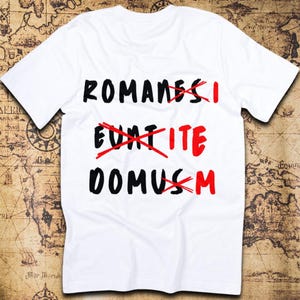 Pode incluir: Uma t-shirt branca de manga curta com o texto preto ROMANES EUNT DOMUS. As letras I, ITE e M estão escritas a vermelho, com marcas de X vermelhas a riscar as letras pretas originais S, EUNT e US respetivamente.