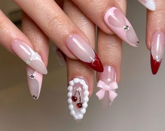 May include: A set of long, almond-shaped acrylic nails. The nails feature a variety of designs, including pink and white gradients, heart shapes, bows, and cherry accents. Some nails have rhinestones for added sparkle.