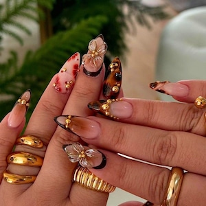 Könnte beinhalten: Nahaufnahme von Händen mit langen, mandelförmigen Nägeln, die verschiedene Nagelkunstdesigns zeigen. Einige Nägel haben ein Schildpattmuster, andere ein Blumenmuster und einige einen Verlaufseffekt. Die Nägel sind mit kleinen goldenen Perlen verziert. Die Hände tragen auch mehrere goldene Ringe.