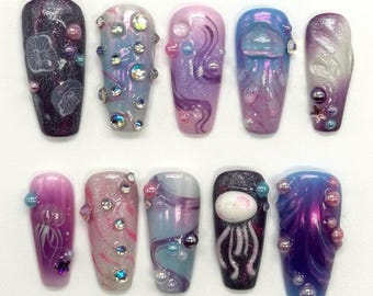 May include: Ten long, oval-shaped fake nails with a variety of designs. The nails feature a purple, blue, and white color scheme with a variety of embellishments, including pearls, glitter, and 3D jellyfish designs.