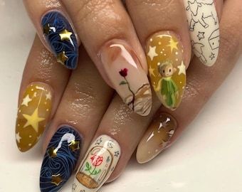 May include: Close-up of a hand with several almond-shaped nails painted with different designs inspired by The Little Prince. The designs include a rose in a glass dome, a yellow star, a blue sky with stars, and a drawing of The Little Prince.